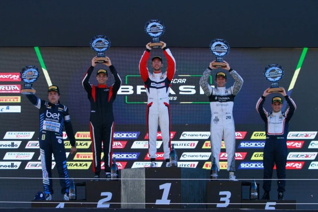 Vencedores no pódio da NASCAR Brasil Series levantando seus troféus. Gabriel Casagrande está no centro, vestindo um macacão branco e vermelho da Militec1, cercado pelos outros pilotos nas posições de segundo e terceiro lugar, todos segurando troféus e celebrando suas conquistas.