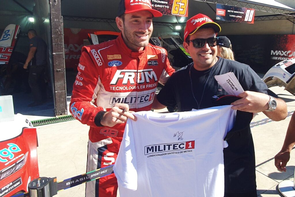 Piloto Vitor Genz, vestindo um macacão vermelho da Militec1, sorri ao lado de um fã que segura uma camiseta branca da Militec1. A cena acontece na área dos boxes, com o carro de corrida ao fundo.