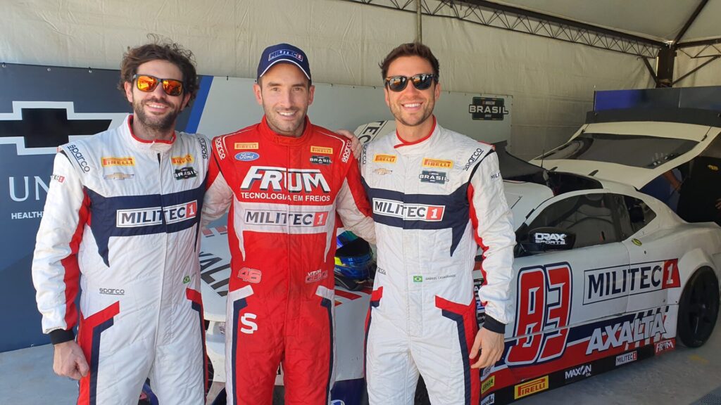 Pilotos Vitor Genz, Gabriel Casagrande e Seid Genz posam para uma foto na frente de um carro de corrida Militec1. Todos estão sorrindo e usando macacões de corrida com as cores e logotipos da Militec1, destacando a camaradagem entre eles.