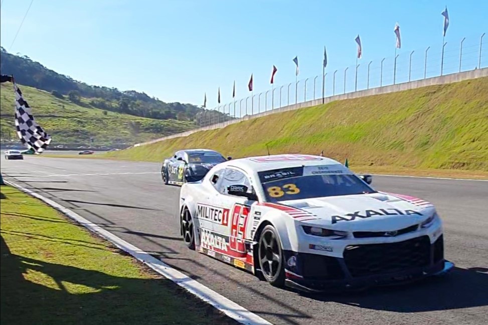 Carro de corrida número 83 da Militec1 cruza a linha de chegada no Autódromo de Potenza, com a bandeira quadriculada visível no canto da imagem. Outros carros de corrida estão em segundo plano, enquanto o cenário montanhoso complementa a ação da pista.