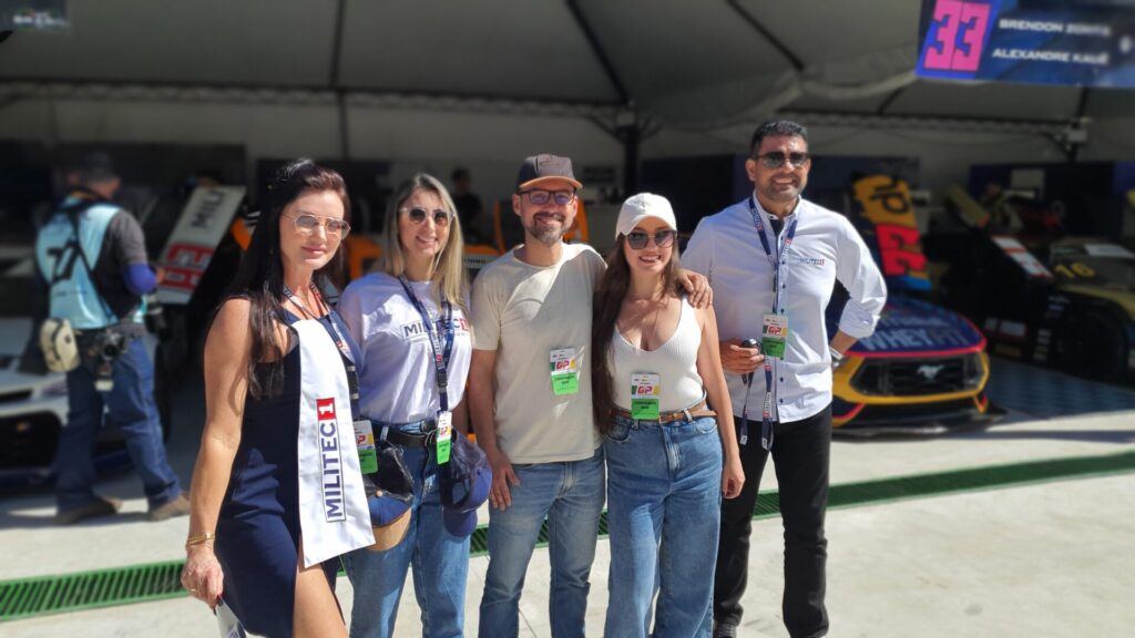 Grupo de seis pessoas, incluindo fãs e membros da equipe da Militec1, posam para uma foto na área dos boxes. Todos estão sorrindo, vestindo roupas casuais e camisetas com o logotipo da Militec1, demonstrando a interação entre a equipe e os fãs.