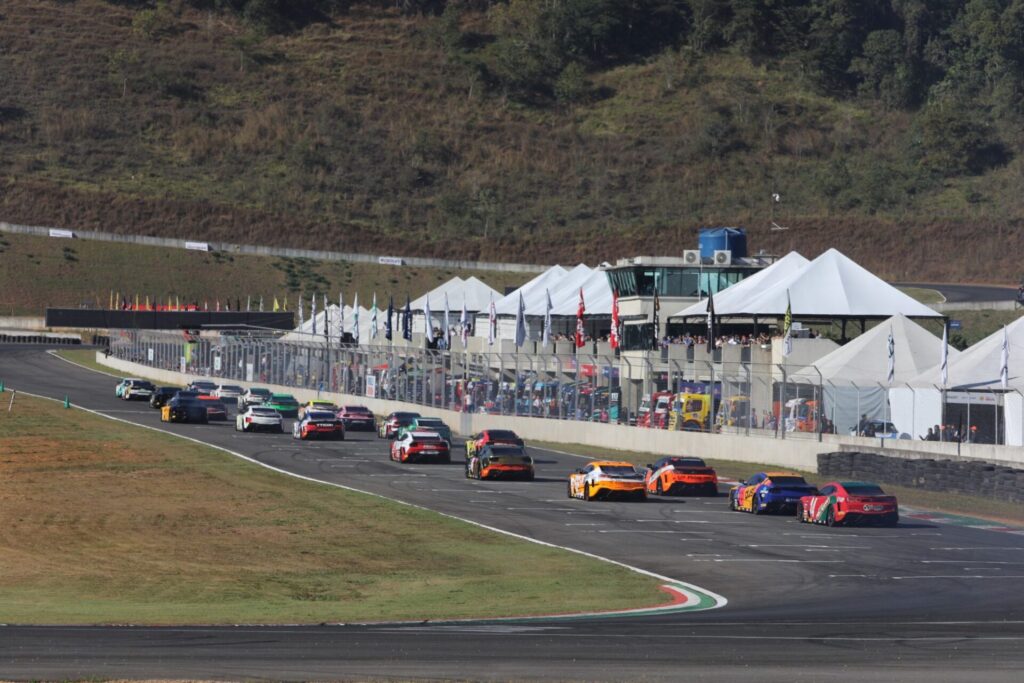 Corrida da NASCAR Brasil Series no Autódromo de Potenza, com vários carros de corrida contornando a curva. Tendas brancas e a área dos boxes são visíveis ao fundo, enquanto a paisagem montanhosa adiciona um belo pano de fundo à cena de alta velocidade.