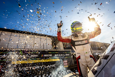 Piloto Gabriel Casagrande, vestindo um macacão de corrida branco e vermelho da Militec1, comemora sua vitória na NASCAR Brasil Series. Ele está no pódio, levantando as mãos, com confetes caindo ao seu redor e o sol brilhando atrás dele. A cena captura a emoção e o triunfo do momento.